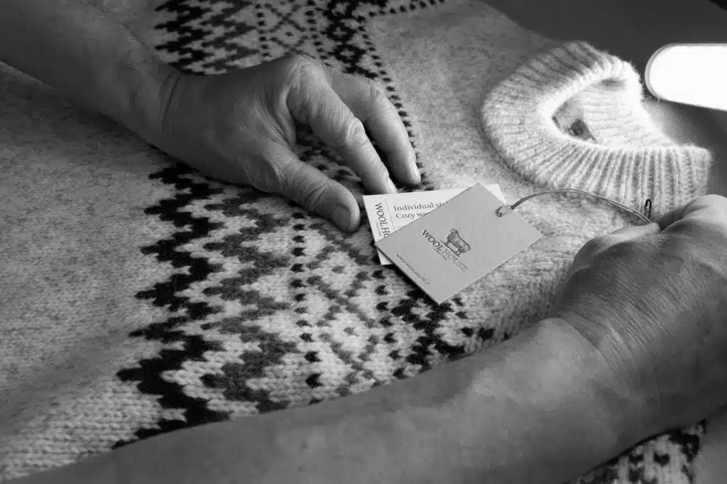 Meistrės rankos prie šiaurietiško rašto megztinio su Wool House etiketėmis
